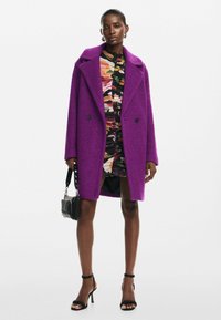 Abrigo morado texturizado con cuello de pico, forma oversize, combinado con un vestido colorido y estampado, bolso negro y tacones con tiras.