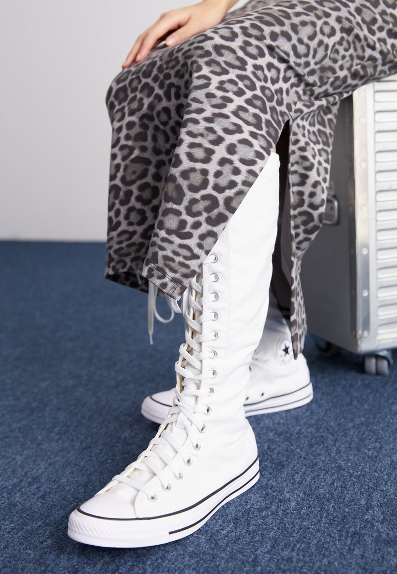 Botas brancas de amarrar com cano alto, feitas de lona, apresentam detalhes em preto. Combinadas com uma saia cinza com estampa de leopardo que tem fendas laterais.