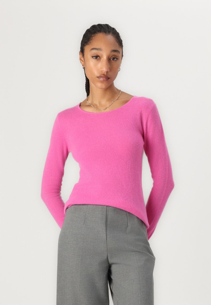 Figurbetontes rosa Kaschmirpullover mit langen Ärmeln und rundem Ausschnitt, kombiniert mit maßgeschneiderten grauen Hosen. Weiche Textur, glatte Oberfläche.