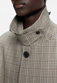 Cappotto scozzese con alto collo, tonalità marrone e beige, tessuto texturizzato e due bottoni neri vicino al collo.