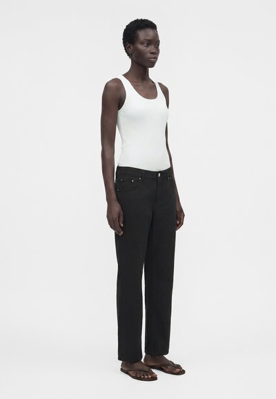 Femme debout portant un haut blanc sans manches, un pantalon noir et des tongs marron foncé sur un fond blanc uni.