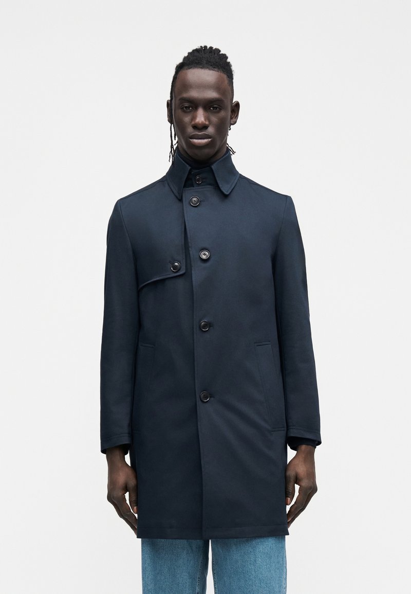 Marineblauer Trenchcoat mit hohem Kragen, Knopfleiste vorne und zwei Seitentaschen. Hergestellt aus glattem Material mit einer strukturierten Silhouette.
