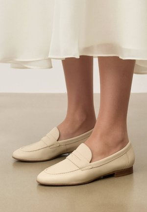 Beige lædermokkasiner med lave træhæle båret med et cremefarvet skørt på en glat beige gulv.