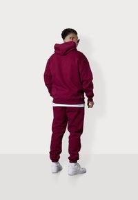 Un sweat à capuche bordeaux et un pantalon de jogging en tissu doux. Le sweat présente une grande poche avant, tandis que le pantalon a des poignets côtelés. Des baskets blanches sont visibles.