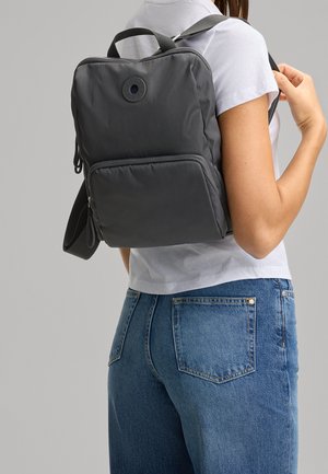 Grauer Nylon-Rucksack mit einer mit Reißverschluss versehenen Fronttasche und verstellbaren Riemen, der ein kreisförmiges Logo an der Oberseite sowie eine glatte Oberfläche aufweist.