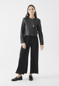 Giubbotto in pelle nera cropped con zip diagonali, abbinato a pantaloni larghi neri a pieghe e mocassini neri. Design semplice e moderno.