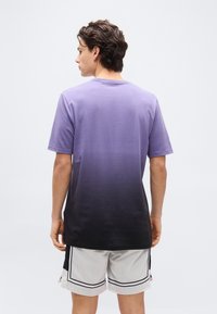 Άντρας με καστανά μαλλιά, που φοράει μπλουζάκι με διαβάθμιση από μοβ σε μαύρο και λευκά σορτς από δίχτυ, στέκεται γυρισμένος προς τα πίσω μπροστά σε απλό φόντο.