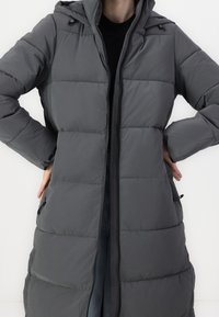 Grijze donsjas met een lang model, verticale doorgestikte structuur, zakken met rits en een verstelbare capuchon. Binnenvoering zichtbaar bij de opening.