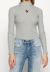Ljust grå ribbad långärmad topp med hög hals, prydd med röda rosor broderade på bröstet; kombinerad med blå denimjeans.