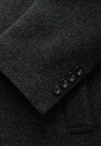 Manga de un blazer de lana gris oscuro con textura de espiga y cuatro botones negros decorativos en el puño.