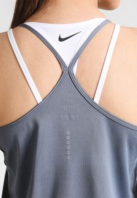 Sportowy top na ramiączkach Nike, wykonany z szarej siateczki z białymi ramiączkami. Posiada krzyżujące się paski na plecach oraz logo w górnej części.