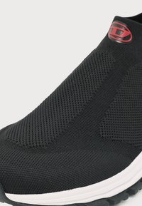 Κοντινό πλάνο ενός μαύρου πλεκτού sneaker χωρίς κορδόνια με λευκή σόλα και κόκκινο λογότυπο στο μανσέτα του αστραγάλου.
