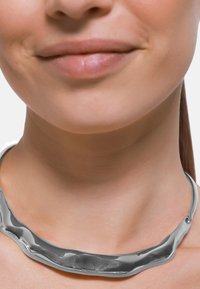 Collana in argento con un design ondulato e asimmetrico, caratterizzata da una superficie liscia. La lunghezza si avvolge comodamente attorno al collo. Hardware minimalista.