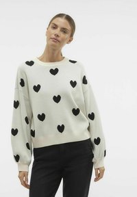 Pull blanc avec motif de cœur noir, col rond et manches longues. Tissu doux, coupe décontractée et poignets côtelés.