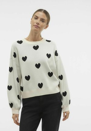 White sweater with black heart pattern, round neckline, and long sleeves. Soft texture, relaxed fit, and ribbed cuffs at the wrists.