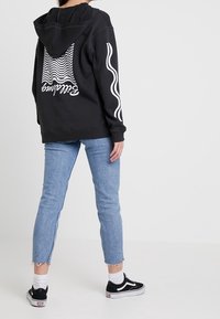 Sudadera negra con patrones ondulados blancos, combinada con jeans de color azul claro con dobladillos deshilachados y zapatillas negras con detalles blancos.