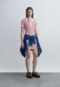 Femeie purtând o rochie cu dungi roșii și albe, cu o jachetă albastră legată în jurul taliei și pantofi maro cu platformă și șireturi.