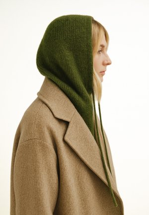 Beiger Wollmantel mit einem strukturierten Kragen, kombiniert mit einer grünen, gestrickten Kapuze mit langen Bändern, aus der Seitenansicht gezeigt.