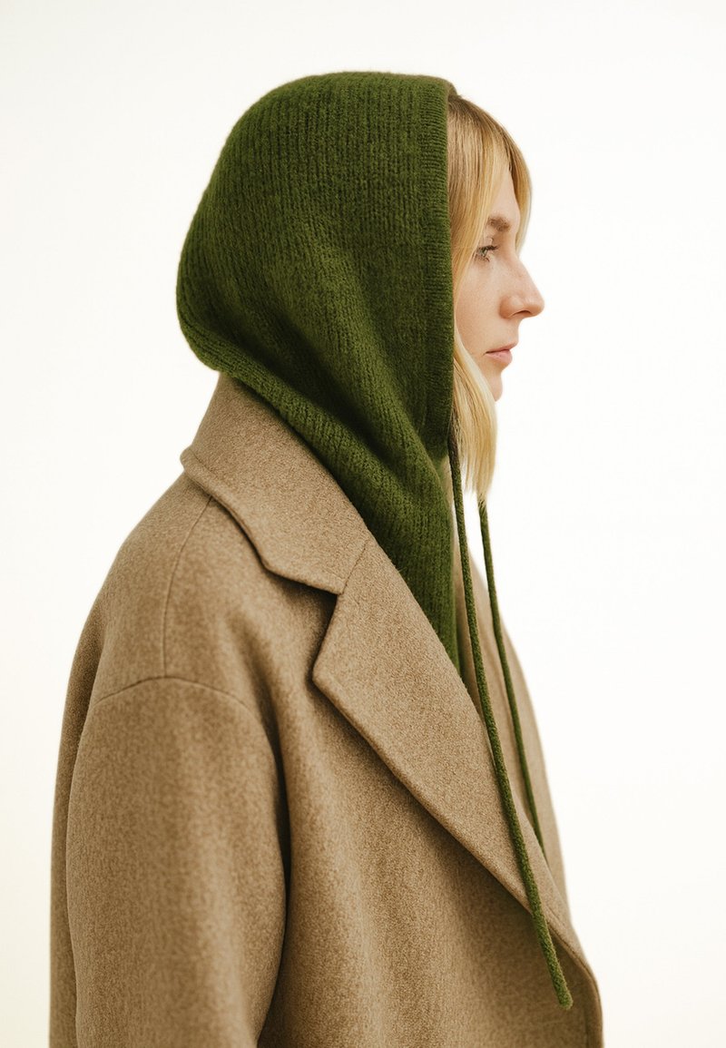 Beiger Wollmantel mit einem strukturierten Kragen, kombiniert mit einer grünen, gestrickten Kapuze mit langen Bändern, aus der Seitenansicht gezeigt.