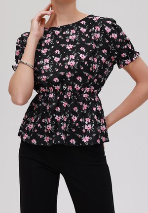Vrouw draagt een zwart T-shirt met korte mouwen met roze bloemenprint en elastische taille, gecombineerd met een zwarte broek, zilveren armband om de linkerpols.