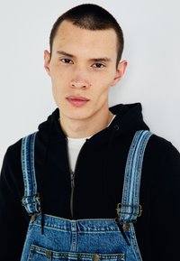 Denim overalls with adjustable straps and metal hardware, worn over a black hoodie. Features a light blue wash and front pocket detailing.