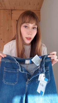 Mladi žena s dugom kosom drži par novih plavih traperica pokazujući prednju stranu s pričvršćenim oznakama u unutrašnjosti.