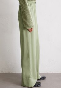 Light green wide-leg trousers with a smooth texture and elastic waistband, featuring side pockets and a subtle sheen. Paired with black flat shoes.