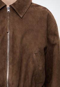 Veste en daim marron avec grand col, fermeture éclair argentée et poche latérale verticale visible sur un côté.