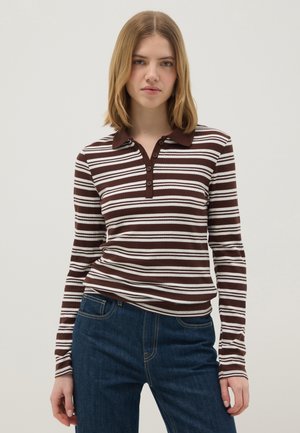 Jeune femme portant un polo à manches longues rayé marron et blanc et un jean bleu foncé, posant devant un fond clair uni.