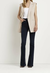 Beige väst med ränder och en knapp, kombinerad med mörka utsvängda jeans. Västen har en skräddarsydd passform och är utan ärmar.