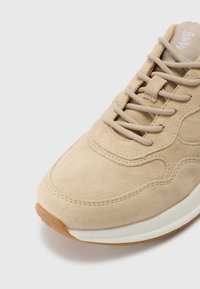 Beige Wildleder-Sneaker mit strukturiertem Obermaterial, Schnürsystem, gepolsterter Ferse und einer weißen Schaum-Mittelsohle. Sichtbare sandfarbene Gummiaußensohle.