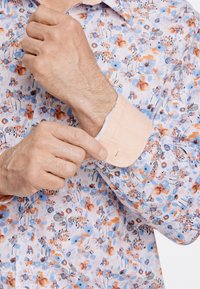Chemise à motif floral dans des couleurs pastel, avec un col orange clair, un tissu texturé, et un gros plan sur le poignet en train d'être ajusté.