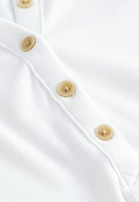 Wit ribmateriaal met een rij grote, ronde beige knopen met zichtbare stiksels, wat een gestructureerde uitstraling geeft.
