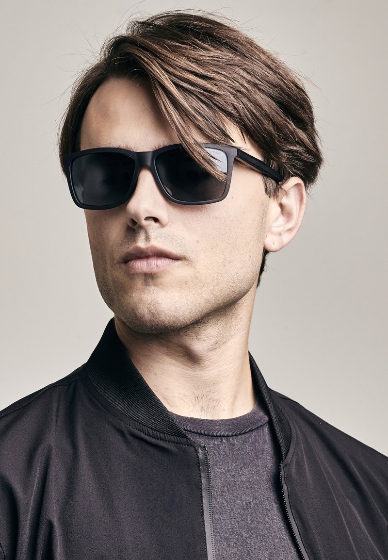 Čierne slnečné okuliare s obdĺžnikovým tvarom, tmavými sklami a elegantnými plastovými rámami, v kombinácii s čiernou bomber bundou cez šedé tričko.