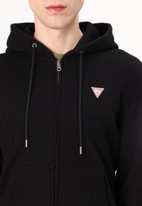 Sweatshirt preto com fecho e ajustável por cordão, apresentando um pequeno logo triangular vermelho e branco no peito, feito de tecido suave.