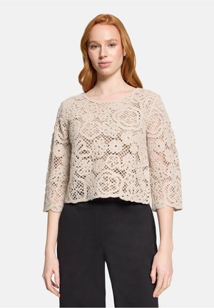 Femme portant un haut en dentelle crochetée fleurie beige avec des manches trois-quarts et un pantalon noir, debout devant un fond uni.