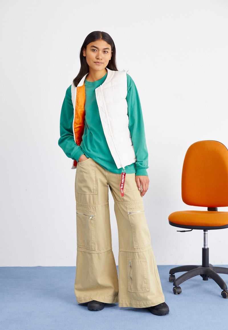 Weißer, plüschiger Weste mit orangefarbenem Innenfutter, grünes Langarmshirt, beigefarbene, weite Cargo-Hose mit Reißverschlüssen, schwarze Schuhe. Oranger Stuhl im Hintergrund.