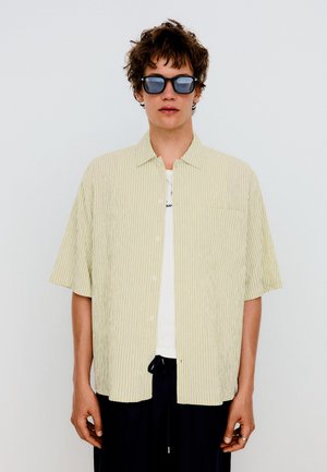 Giovane uomo con capelli ricci indossa occhiali da sole neri, una camicia larga beige chiaro a righe a maniche corte, una canottiera bianca e pantaloni scuri.