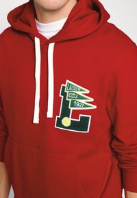 Homme portant un sweat à capuche rouge avec des cordons blancs et un logo Lacoste 1927 vert et jaune avec une balle de tennis sur la poitrine.