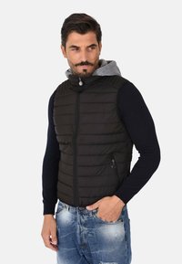 Gilet trapuntato nero con colletto alto, chiusura con zip e tasche. Cappuccio grigio visibile sotto. Indossato sopra una maglietta a maniche lunghe blu navy.