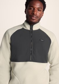 Fleece pullover in light cream with dark gray front panel. Features a high collar and zip closure; textured fabric with soft, brushed finish.