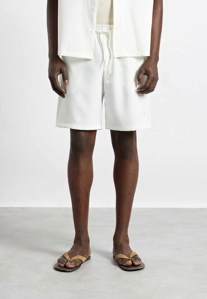 Personne à la peau foncée portant une chemise blanche boutonnée, un short blanc à cordon et des sandales marron type tongs, debout sur un sol gris.