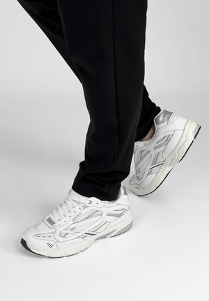 Persoon met zwarte broek en witte mesh sneakers met het label "Dutch D", staand met één voet iets omhoog tegen een effen lichte achtergrond.