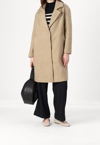 Béžový kabát s texturou, klopovým límcem, bočními kapsami a přímým střihem, spárovaný s černými širokými kalhotami a světlými balerínami. Drží černou kabelku.