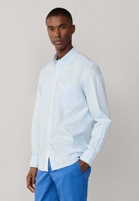 Homme portant une chemise à rayures bleu clair à boutons et un pantalon bleu cobalt, debout avec une main dans la poche, sur un fond uni.