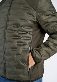 Olivgrön puffjacka med kviltat mönster, dragkedja fram, sidofickor och texturerade ärmar med nätinslag.
