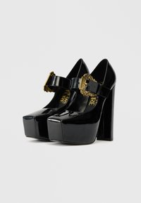 Zapatos de tacón tipo plataforma en charol negro con punta cuadrada, un gran tacón bloque y una hebilla decorativa dorada en una correa ajustable.
