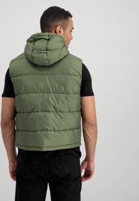 Padded bezrukávový vest v olivovej zelenej farbe s kapucňou, ktorý má horizontálne šité sekcie a hladký, vodeodolný povrch.