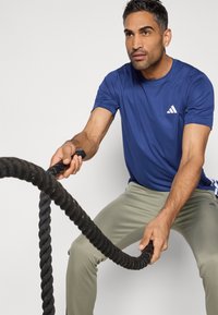 Zils sporta T-krekls ar baltu logo, kopā ar gaismi zaļām biksēm, persona, kas satver melnu cīņas virvi, koncentrēta izteiksme, neitrāla fona.