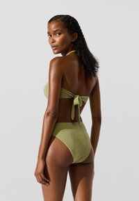 Zaļš spīdīgs bikini komplekts ar bandeau topu, kam ir aiztaisāms aizmugurē, un augsta vidukļa apakšdaļu, kas demonstrē gludu, teksturētu virsmu.